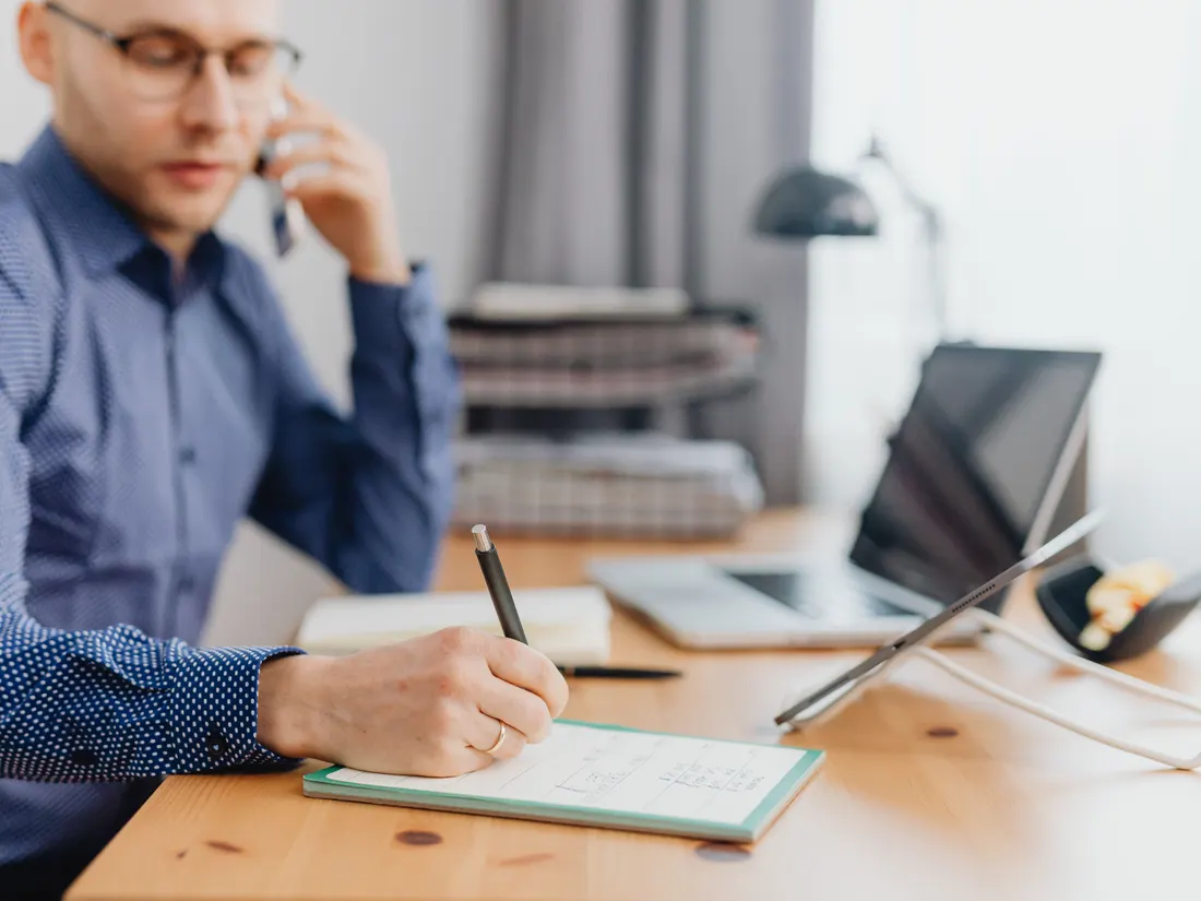A person taking notes at a desk while on the phone, with a laptop and tablet beside them