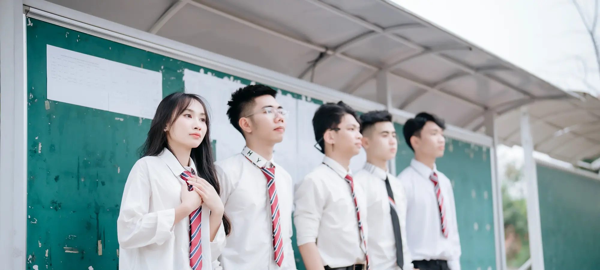 Secondary-school students in uniform standing along a covered walkway beside a classroom noticeboard