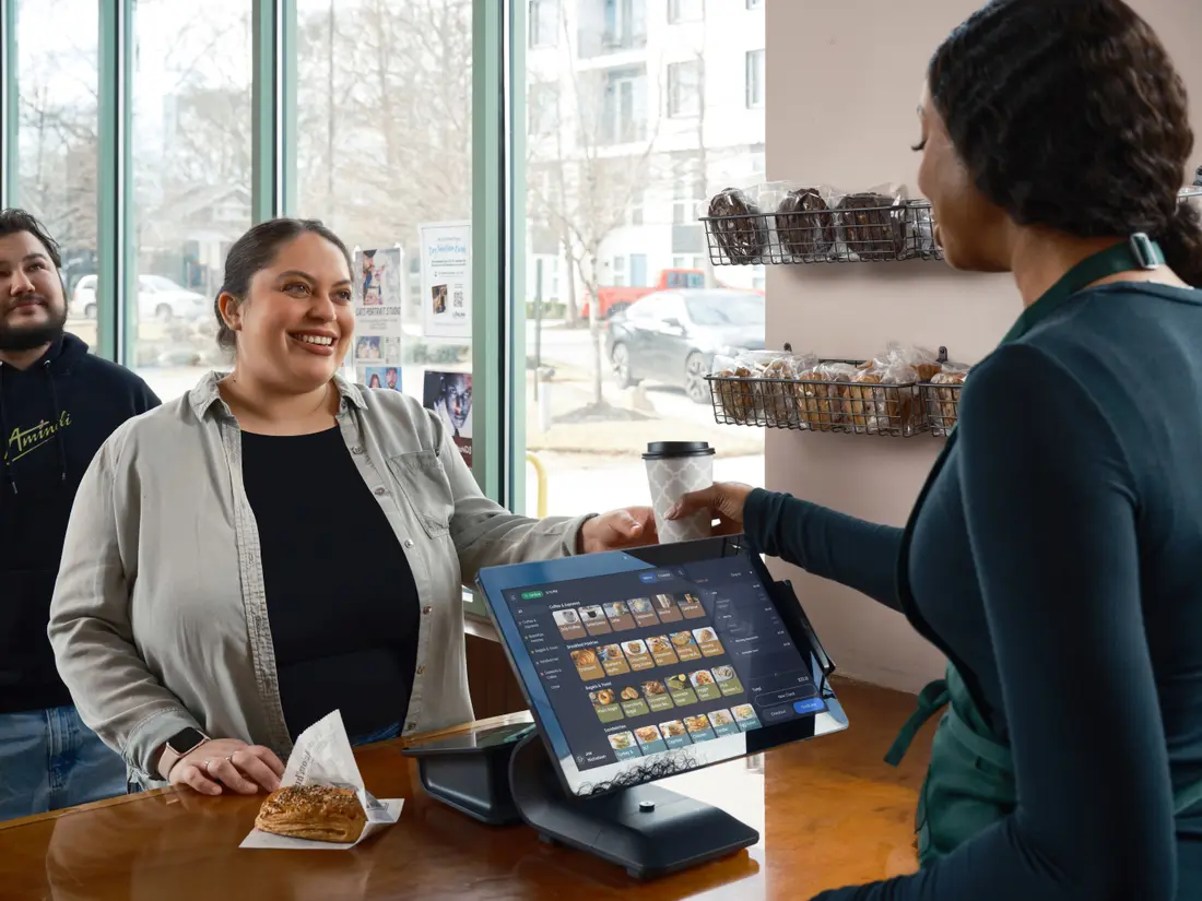 Two staff members serving a customer coffee at a café counter with a till